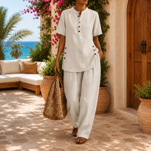 May include: A light beige short-sleeved shirt with a mandarin collar and a partial button-down front, paired with matching loose-fitting trousers. The shirt features decorative buttons along the side seams and is worn with a woven brown tote bag.