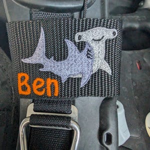 Pode incluir: Patch de tecido preto com bordados de tubarões-martelo e outros tubarões. O nome "Ben" é bordado em laranja. O patch é preso a uma alça preta com fivela e argola em D de metal.