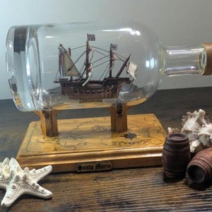 May include: A detailed model ship inside a clear glass bottle, mounted on a wooden base with a map design. The bottle has a wooden stopper. Decorative elements include a starfish and small wooden barrels.