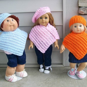 May include: Three dolls wearing crocheted ponchos and hats. The doll on the left is wearing a light blue poncho and hat, the doll in the middle is wearing a pink poncho and hat, and the doll on the right is wearing an orange poncho and hat.