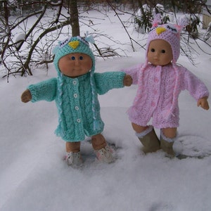 May include: Two dolls in knitted owl-themed outfits stand in the snow. One doll wears a light blue outfit with a matching hat, the other a pink outfit. Both dolls have brown shoes and are surrounded by snow. The outfits are handmade.