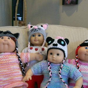 May include: Four dolls wearing handmade crocheted hats. The hats are designed as a raccoon, cat, panda, and ladybug. The dolls are wearing knitted sweaters with braided yarn pigtails.