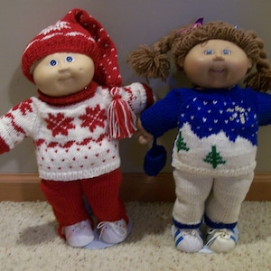 May include: Two Cabbage Patch Kids dolls wearing knitted winter outfits. The doll on the left is wearing a red and white sweater with a snowflake pattern, red pants, and a red and white hat. The doll on the right is wearing a blue and white sweater with a snowflake pattern, white pants, and a blue hat.