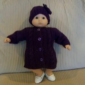 May include: A doll dressed in a knitted purple coat, hat, and shoes. The coat fastens with buttons, and the hat features a bow. The doll is positioned on a beige cushion.
