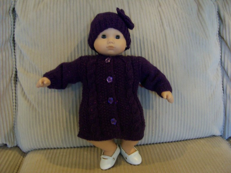May include: A doll dressed in a knitted purple coat, hat, and shoes. The coat fastens with buttons, and the hat features a bow. The doll is positioned on a beige cushion.