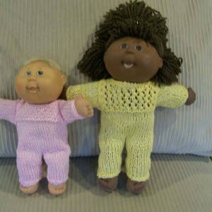 Puede incluir: Dos muñecas Cabbage Patch de punto con atuendos hechos a mano. Una muñeca tiene el pelo castaño y lleva un atuendo de punto amarillo. La otra muñeca tiene el pelo rubio y lleva un atuendo de punto rosa.