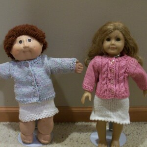 May include: Two dolls are displayed, each wearing a knitted outfit. One doll has brown hair and a blue and white sweater with a white skirt. The other doll has blonde hair and a pink sweater with a white skirt. Both dolls are standing on clear stands.