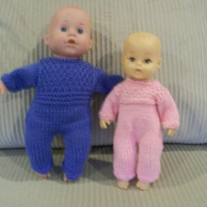 May include: Two baby dolls wearing knitted outfits. The doll on the left is wearing a purple knitted jumpsuit. The doll on the right is wearing a pink knitted jumpsuit.