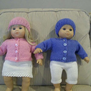 May include: Two dolls wearing handmade knitted outfits. One doll is dressed in a pink sweater, skirt, and hat. The other doll is wearing a blue sweater, hat, and white pants. Both outfits have button closures.