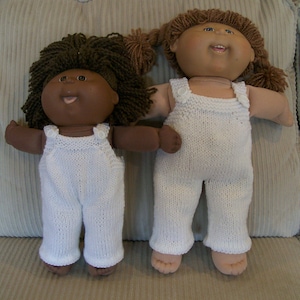 Könnte beinhalten: Zwei Cabbage Patch Kids Puppen, die weiße gestrickte Latzhosen tragen. Die Puppe links hat braune Haut und braunes Haar. Die Puppe rechts hat helle Haut und braunes Haar.