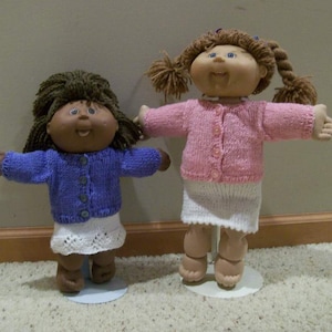 Könnte beinhalten: Zwei Cabbage Patch Kids Puppen, die gestrickte Pullover und Röcke tragen. Die Puppe links trägt einen lila Pullover und einen weißen Rock mit Spitzenbesatz. Die Puppe rechts trägt einen rosa Pullover und einen weißen Rock.