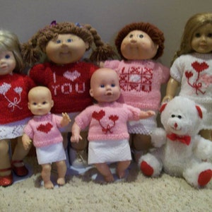 May include: A group of dolls and a teddy bear, all dressed in Valentine's Day themed outfits. The dolls wear red, pink, and white sweaters with heart designs. The teddy bear is white with red accents and a bow tie.
