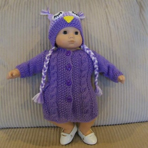 May include: A doll dressed in a purple knitted owl outfit, featuring a hat with owl eyes and a button-down sweater dress. The doll is wearing white shoes and has two white and purple braids. The outfit is a solid purple colour.