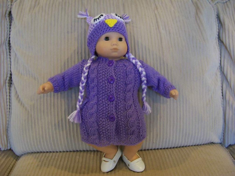 May include: A doll dressed in a purple knitted owl outfit, featuring a hat with owl eyes and a button-down sweater dress. The doll is wearing white shoes and has two white and purple braids. The outfit is a solid purple colour.