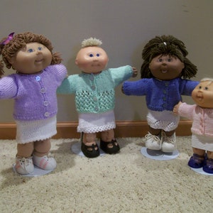 Könnte beinhalten: Vier Cabbage Patch Kids Puppen, die gestrickte Cardigans und Röcke tragen. Die Puppen stehen auf einem hellbraunen Teppich. Die Puppen tragen Cardigans in verschiedenen Farben: lila, hellgrün, blau und rosa.