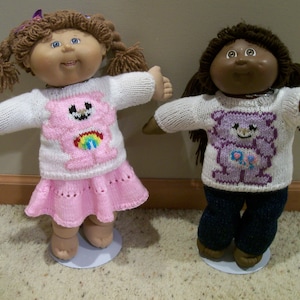 Könnte beinhalten: Zwei Cabbage Patch Kids Puppen, die gestrickte Pullover mit Care Bear-Motiven tragen. Die Puppe links trägt einen rosa Pullover mit einem rosa Care Bear und einen rosa Rock. Die Puppe rechts trägt einen lila Pullover mit einem lila Care Bear.