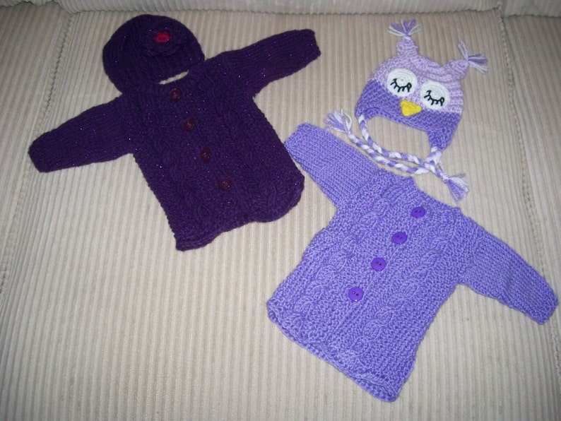 May include: A collection of hand-knitted baby clothes in purple hues. The set features a hooded cardigan with buttons, a matching hat, and an owl-shaped hat. The cardigans have a cable knit pattern. The owl hat has white and purple braided ties.