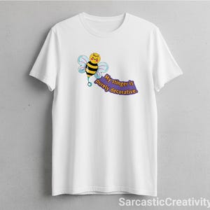 May include: White t-shirt featuring a cartoon bee with black and yellow stripes, blue wings, and a smiling face. The text "My stinger is purely decorative" is on a purple banner. The shirt is a casual wear item.