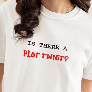 Puede incluir: Camiseta blanca con la frase "IS THERE A PLOT TWIST?" en letras negras y rojas. La camiseta tiene cuello redondo y mangas cortas. La tela parece ser una mezcla de algodón suave.