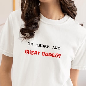 May include: White t-shirt with the text "IS THERE ANY CHEAT CODES?" in black and red. The shirt has a crew neck and short sleeves. The text is centered on the front of the shirt.