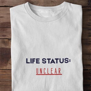 以下が含まれることがあります： 「LIFE STATUS: UNCLEAR」の文字が紺色と赤色でプリントされた白いTシャツ。木製の表面にきれいに折りたたまれています。テキストはシャツの前面の中央に配置されています。