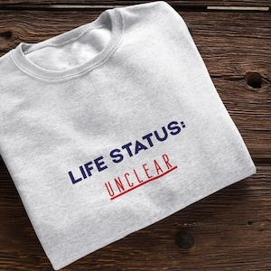 以下が含まれることがあります： 「LIFE STATUS: UNCLEAR」の文字がネイビーブルーと赤でプリントされたライトグレーのスウェットシャツ。木製の表面にきれいに折りたたまれています。クルーネックのスウェットシャツはカジュアルなアパレルアイテムです。