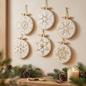May include: Six circular wooden embroidery hoops containing white fabric with intricate snowflake designs stitched in white and gold thread. Each hoop features a screw-top tension mechanism and is suspended by a thin string.