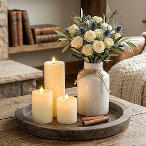 May include: A rustic wooden coffee table displays a decorative arrangement. It features three lit ivory candles, a white vase with roses and thistle, and a wooden tray with cinnamon sticks. A cozy living room setting.