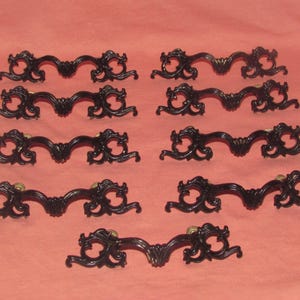 May include: A collection of ten black ornate drawer pulls. Each pull features a curved handle with decorative scrollwork and a central floral design. The pulls are arranged on a peach-colored surface.