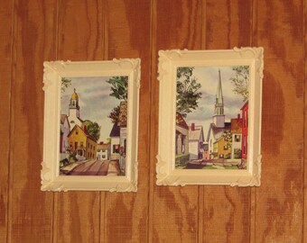 May include: Two rectangular wall art pieces in cream-colored ornate plastic frames. Each print depicts a colorful town street scene with houses, trees, and a church steeple under a cloudy sky. The artwork features yellow, red, and white buildings.