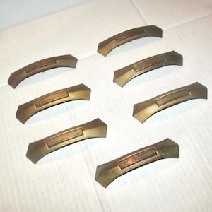 May include: A set of six matching brass-toned metal drawer pulls arranged in two columns. Each handle features a curved, arching design with a rectangular center panel and pointed, angular ends, displaying a weathered, aged metallic finish.