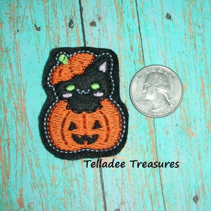 May include: Black cat peeking out of a small orange pumpkin with a green stem. The pumpkin has a jack-o'-lantern face. The image is for size comparison with a US quarter.