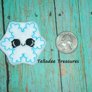 May include: A white felt snowflake with a blue outline and a cute cartoon face. The snowflake is about the size of a US quarter.