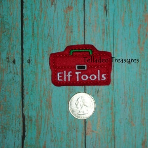 May include: Red felt toolbox with green trim and white embroidery that reads "Elf Tools". The toolbox is on a teal wooden background.