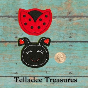 May include: Two felt ladybug appliques, one red with black spots and one black with red hearts and a smiling face. The appliques are shown on a blue wooden background with a US quarter for scale.