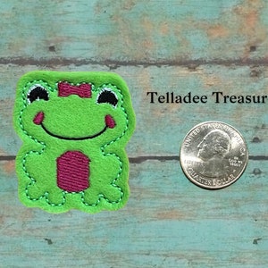 May include: Green felt frog applique with a pink bow, measuring approximately the size of a US quarter.