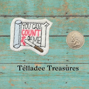 May include: Embroidered patch with a white scroll and the text "You can count on me" in black with "count" in pink. The scroll is rolled up and tied with a brown string. The patch is on a blue wooden background.