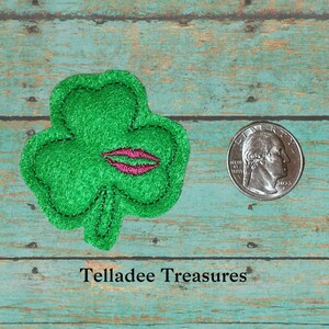 May include: Green felt shamrock with pink lips embroidered on it. The shamrock is about the size of a US quarter.
