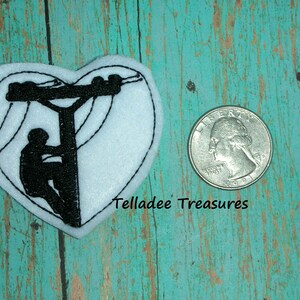 May include: White felt heart-shaped patch with black embroidery of a lineman climbing a utility pole with power lines. The patch is about the size of a quarter.