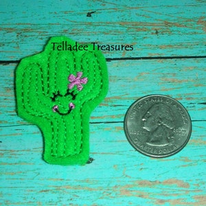 May include: A green felt cactus with a pink flower and a smiling face. The cactus is about the size of a US quarter.