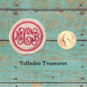 May include: A white fabric circle with a pink border and a monogram embroidered in white. The monogram is "MGB". The circle is about the size of a US quarter.