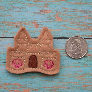 May include: A brown felt sandcastle applique with pink seashells. The sandcastle is about the size of a quarter.