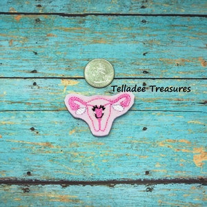 May include: Pink felt applique of a uterus with a smiling face and eyelashes. The uterus is about the size of a US quarter.