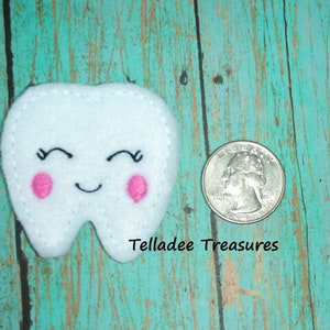 May include: A white felt tooth with a smiling face and pink cheeks. The tooth is about the size of a US quarter.