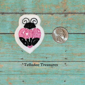 May include: A white felt ladybug applique with black embroidered details. The ladybug's heart is pink and says "Love Bug" in black embroidery. The applique is about the size of a US quarter.