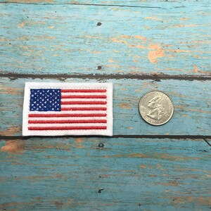 May include: A small embroidered American flag patch on a white background. The flag is red, white, and blue. The patch is about the size of a quarter.