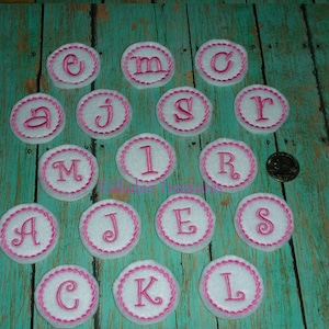 May include: A set of 16 white felt circles with pink stitching around the edges. Each circle has a pink embroidered letter on it. The letters are: A, C, E, I, J, K, L, M, R, S.