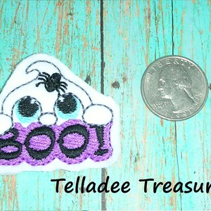 May include: A white felt ghost with black eyes and a spider on top, holding a purple felt banner with the word "BOO!" embroidered in black. The ghost is about the size of a US quarter.