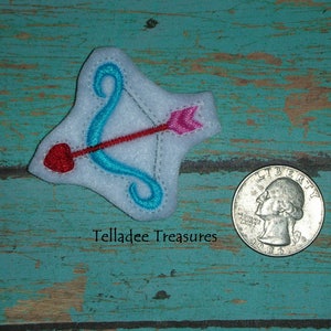 May include: A white felt applique in the shape of Cupid's bow and arrow, with a red heart-shaped arrow tip and a pink arrow head. The bow is turquoise. A quarter is next to the applique. The text "Telladee Treasures" is in the image.