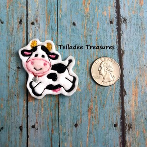 May include: A white and black embroidered cow applique with pink accents and gold horns. The applique is about the size of a US quarter.